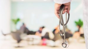 hand holding a stethoscope with a blurred background of patients in a waiting room
